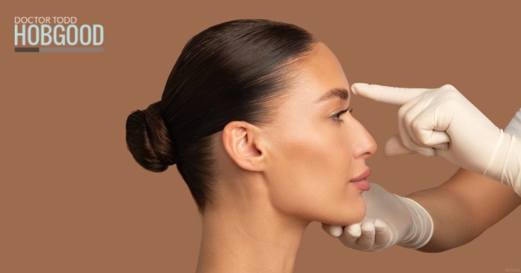 Woman with beautiful nose smiling and touching her face (model) Hobgood Facial Plastic Surgery: Todd Hobgood, MD Logo