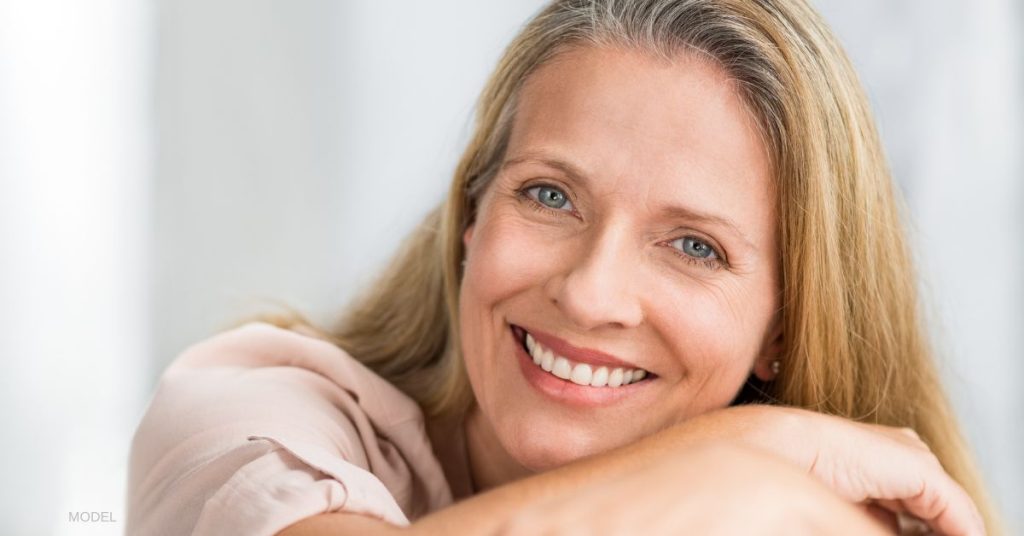 A woman smiling that is resting her head down on the top of her arms. She has blonde/golden colored hair and is wearing a light pink shirt. (model)
